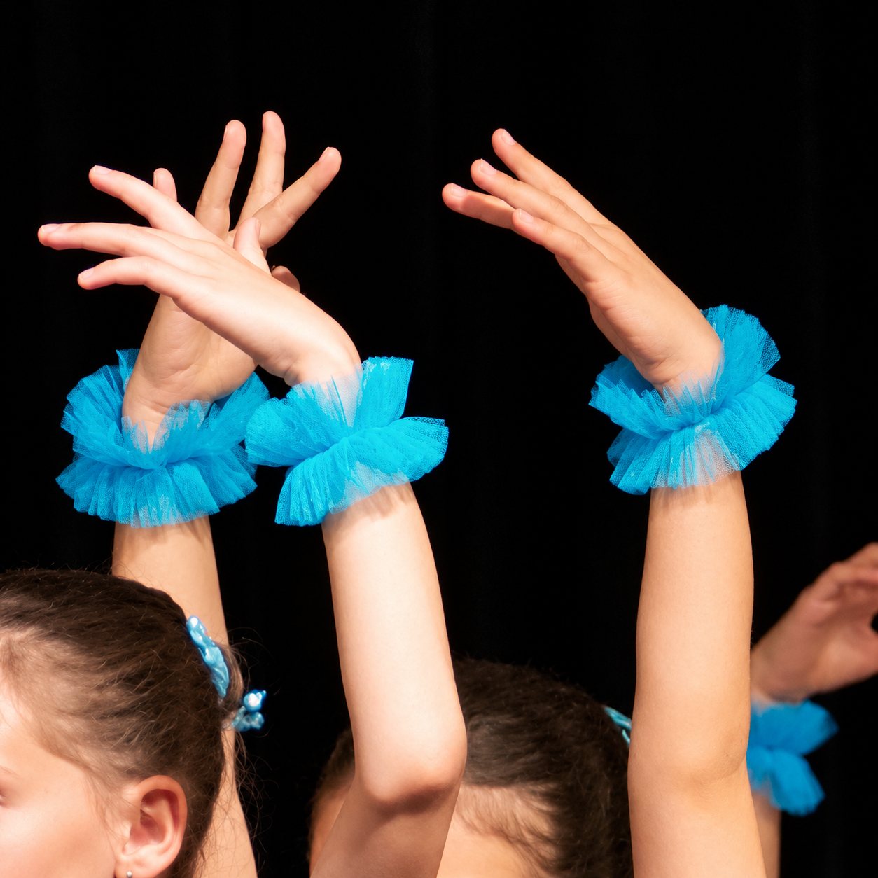 Des bras levés de jeunes danseuses sur scène, ornés de bracelets en tulle bleu turquoise devant un fond noir.
