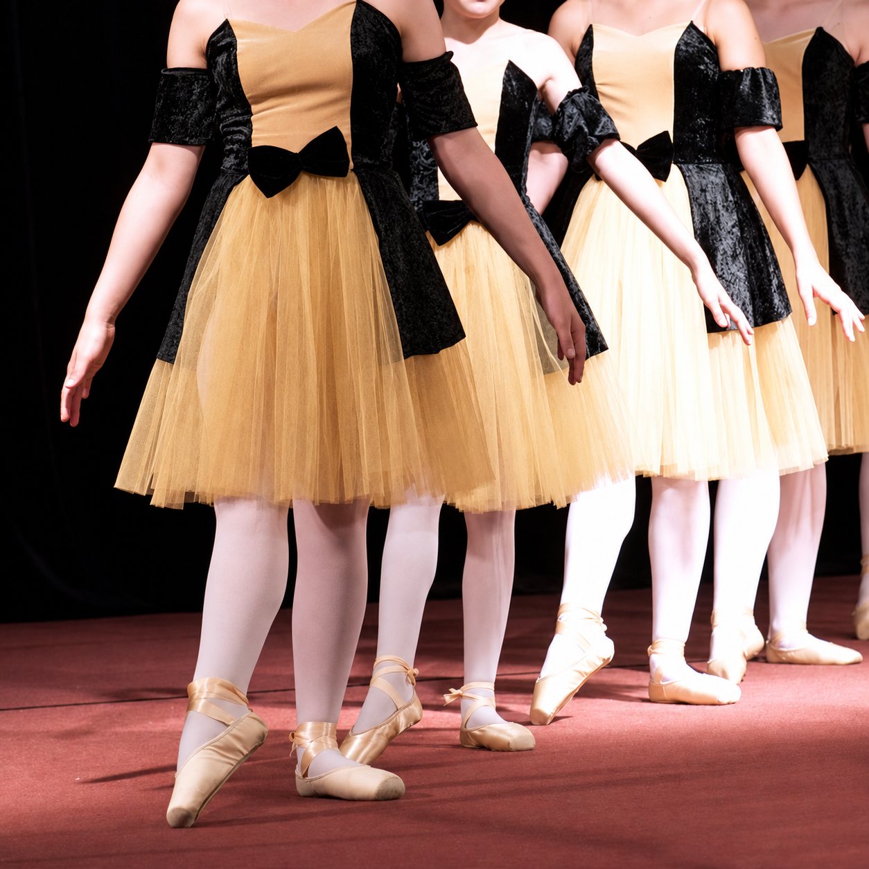 Plusieurs danseuses en pointes et en tutus dorés à corsage de velours noir se tiennent alignées sur une scène au sol rouge.