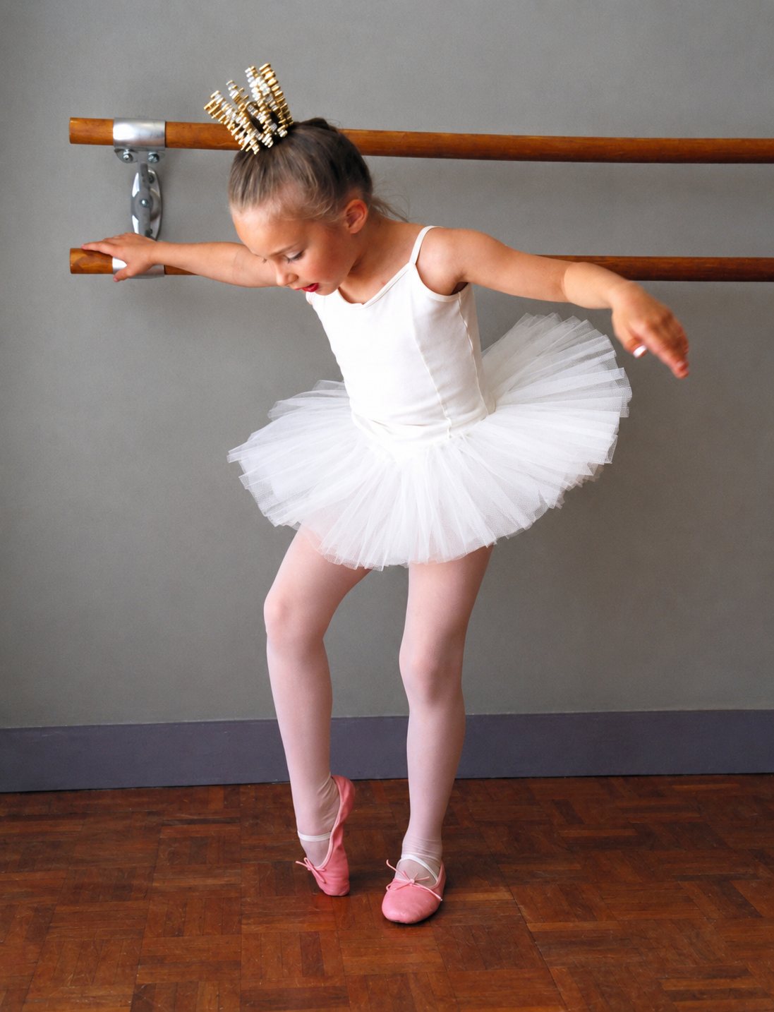 Une petite fille en tutu blanc et chaussons roses, une couronne dans les cheveux, prend la pose près d'une barre de danse dans un studio.