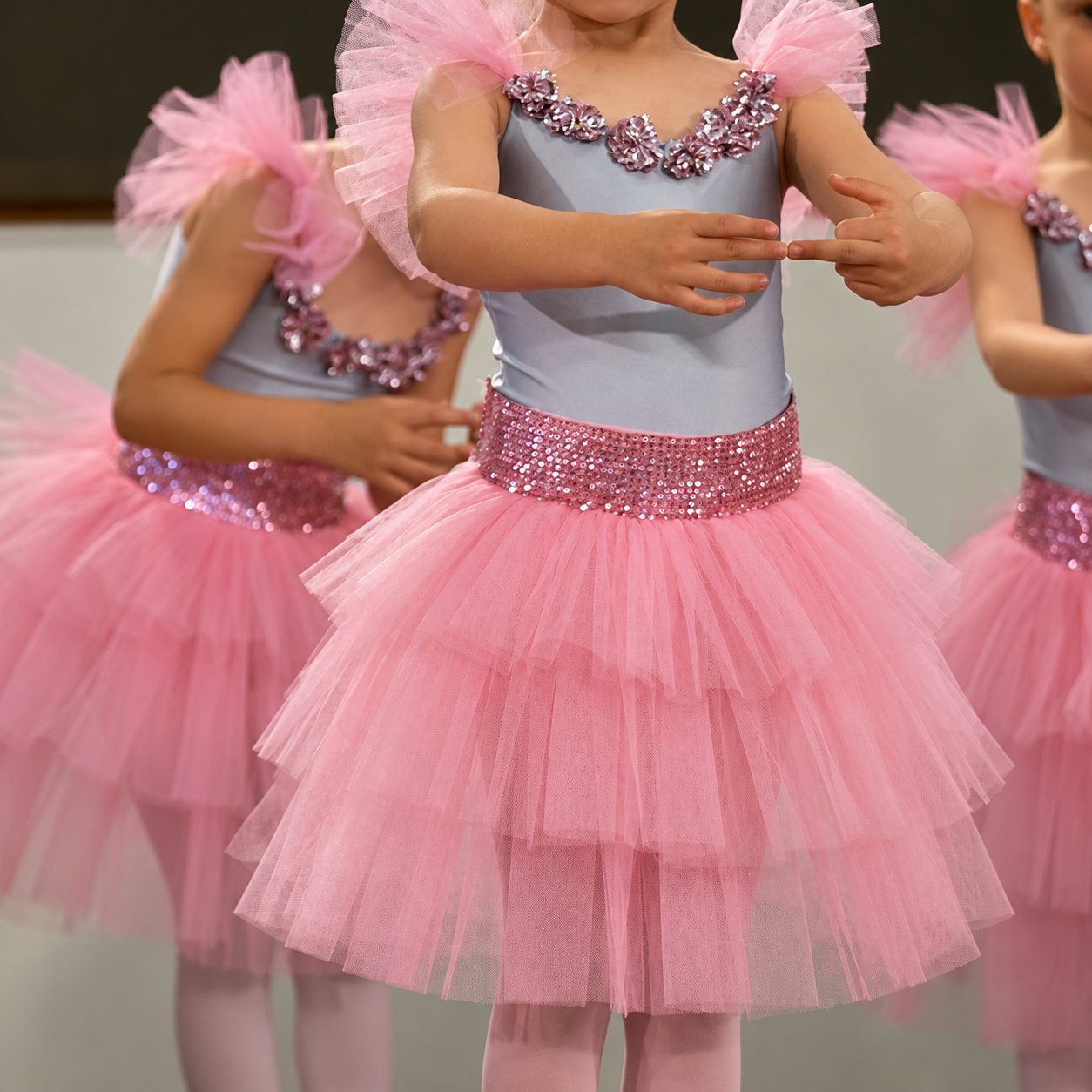 Des petites filles en costume de danse, avec justaucorps gris orné de paillettes et tutu rose en tulle à volants, exécutent un mouvement sur scène.